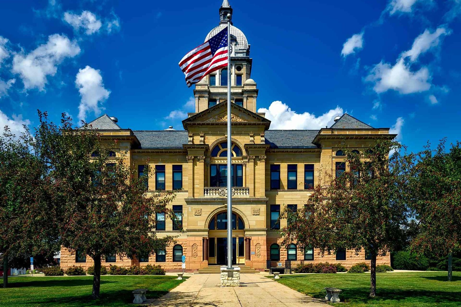 A stone government building with a domed clock tower and a large American flag flying from a pole out front, under a bright blue sky