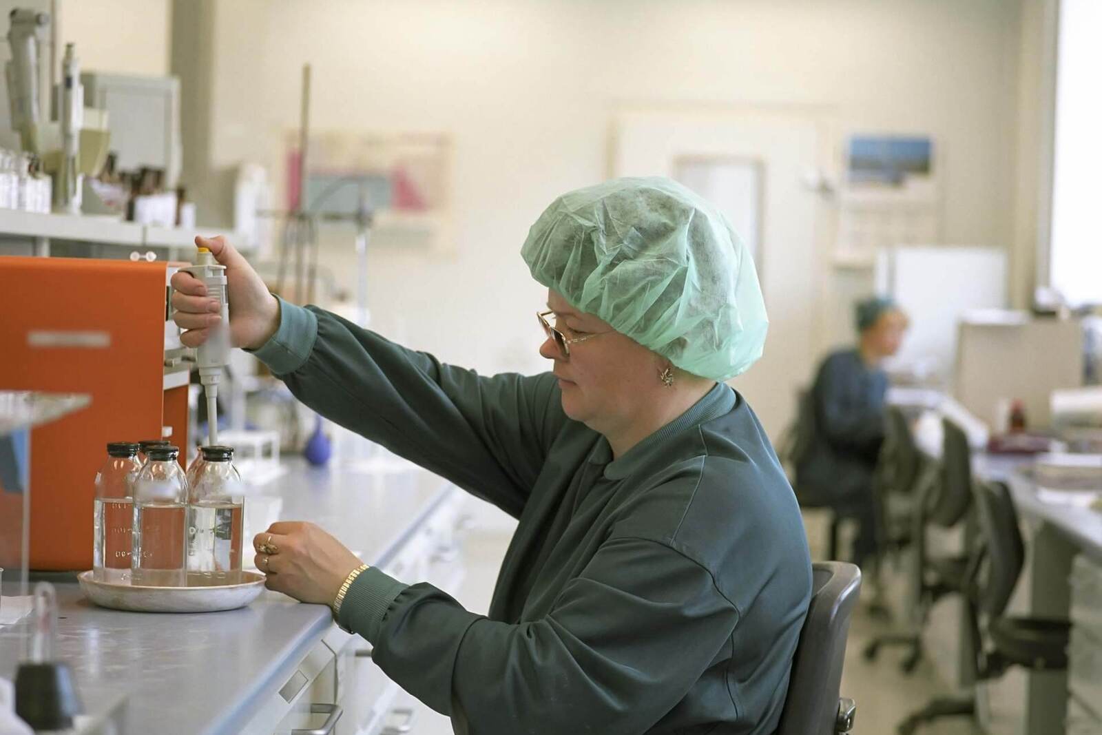 A lab technician in a hairnet and gloves pipetting samples at a bright laboratory bench