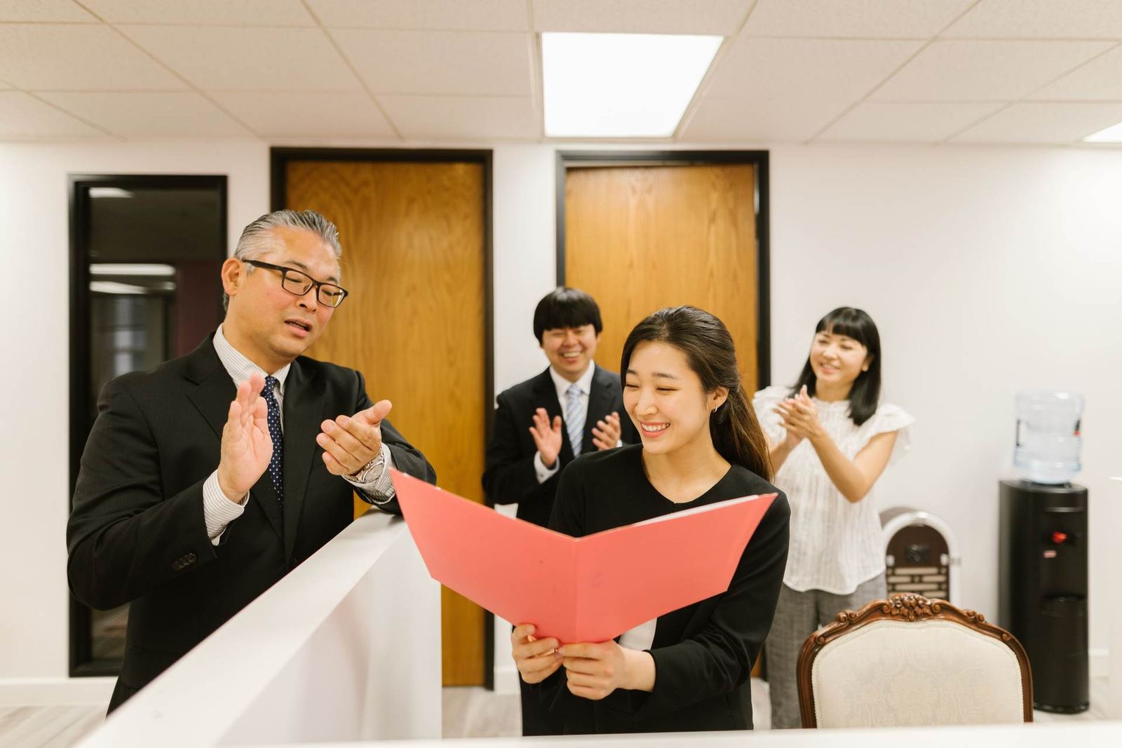 Colleagues applaud as one holds open a red folder in an office setting, marking an official milestone