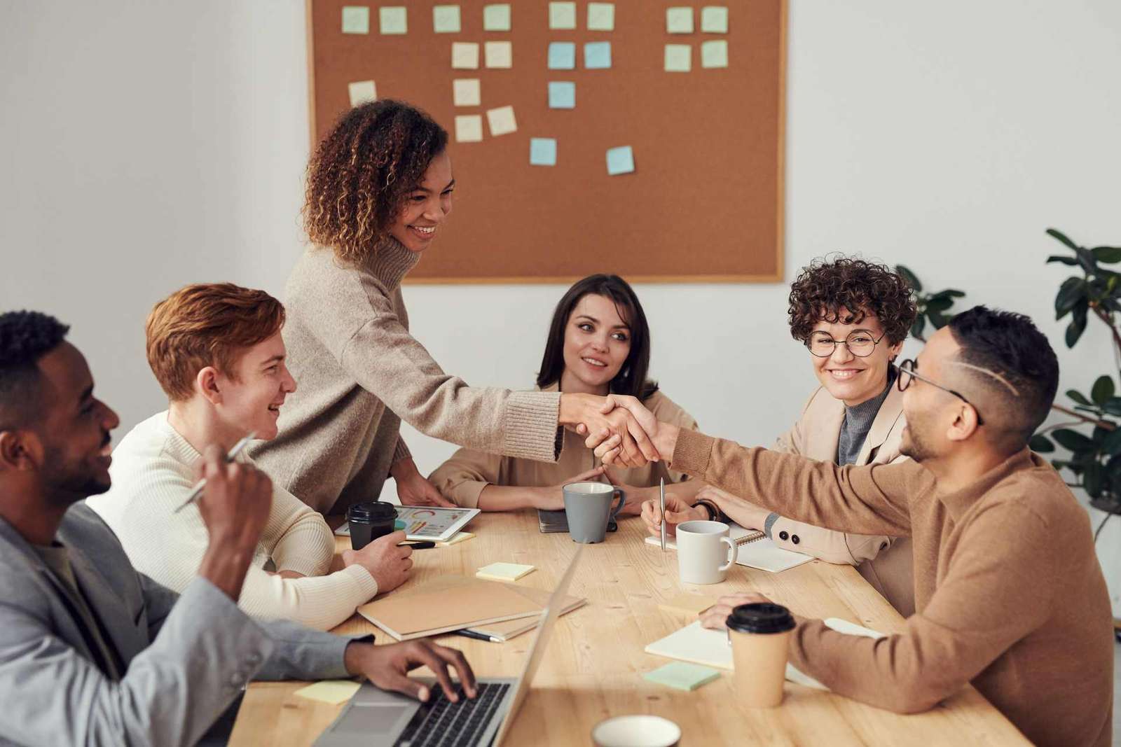 A diverse group of six colleagues around a shared table, two shaking hands and smiling, with notebooks and coffee cups between them