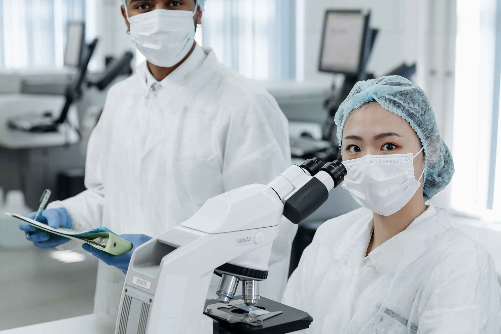Two masked, gowned laboratory researchers in a cleanroom review a sample slide loaded on a light microscope