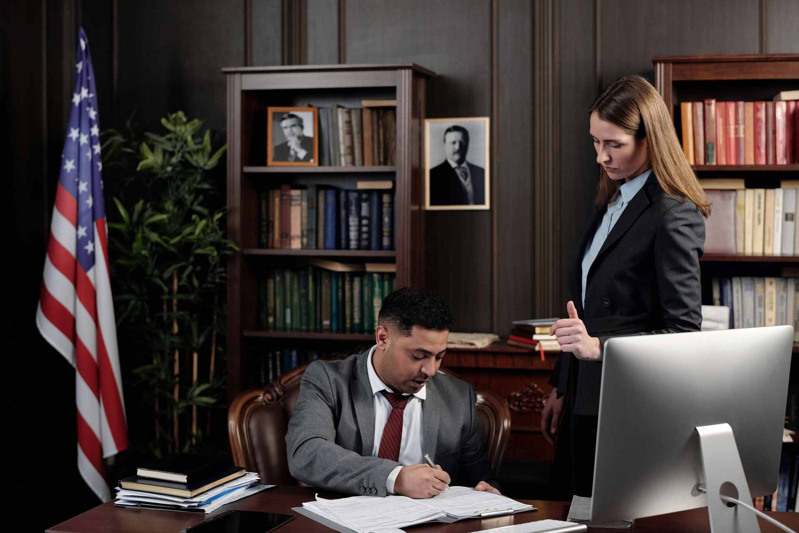 A man in a suit signs paperwork at a desk while a colleague reviews a document beside him in a wood-paneled office with an American flag