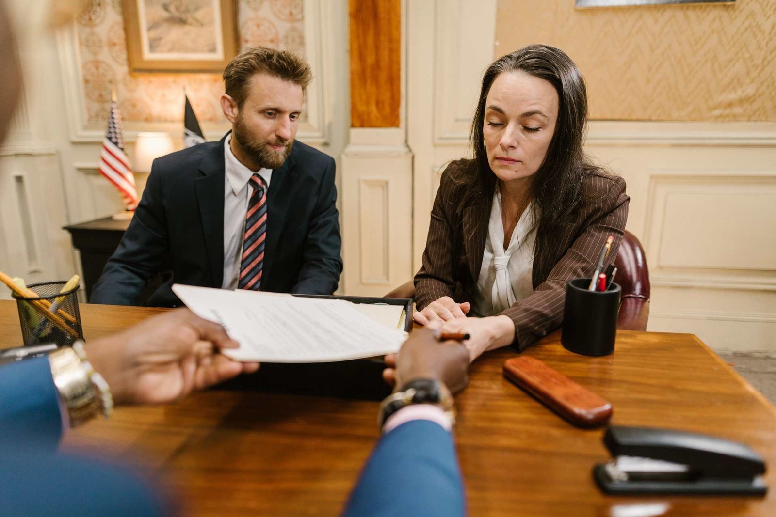 Two officials review a document handed across a desk in a government office, a U.S. flag in the background
