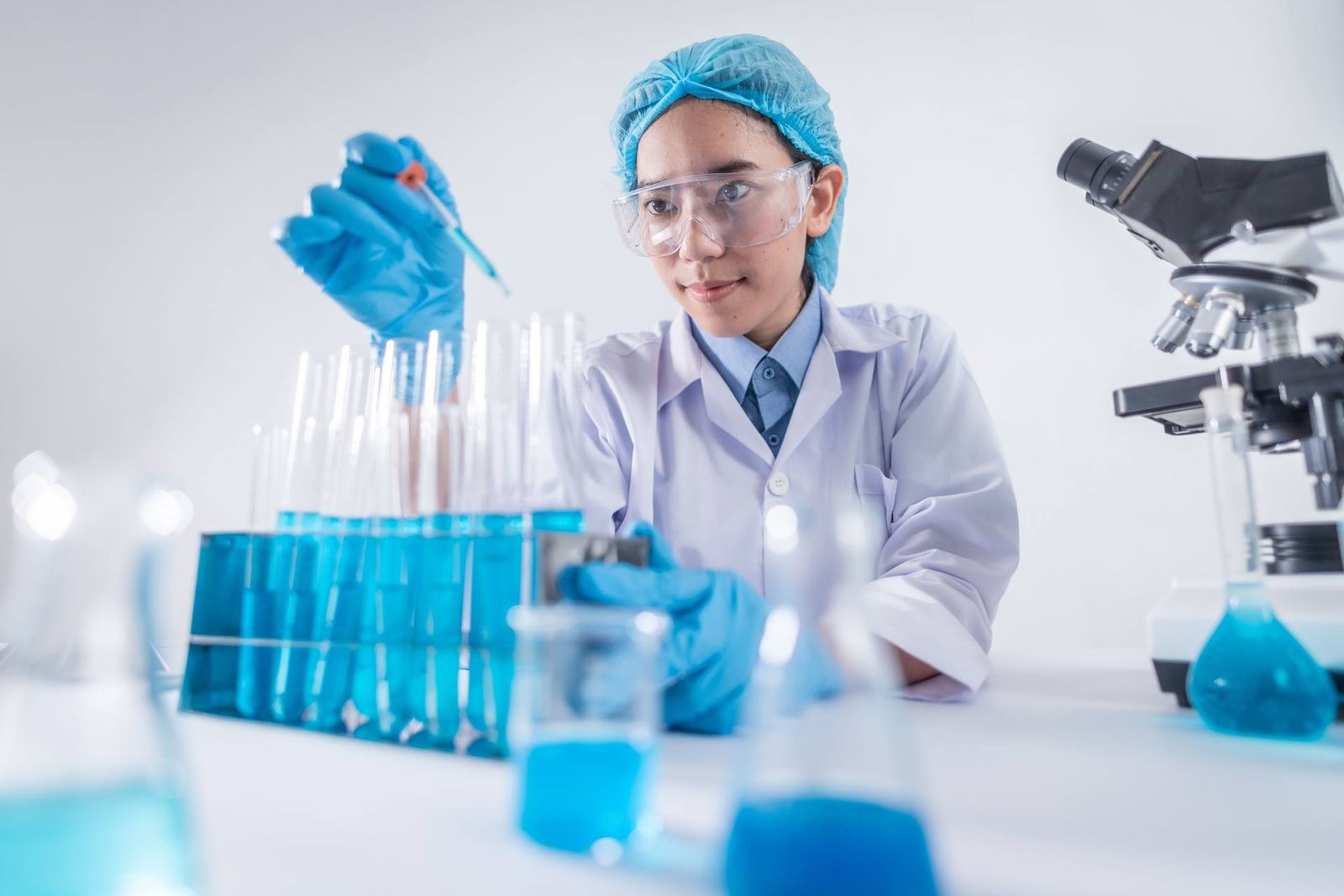 A laboratory scientist in blue nitrile gloves and a hairnet uses a pipette over a rack of test tubes
