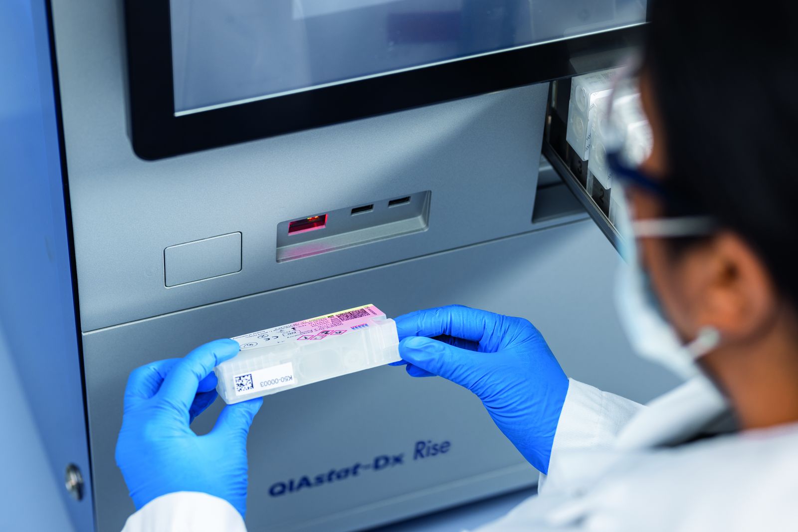 A gloved laboratory technician holding a QIAstat-Dx test cartridge in front of QIAGEN's QIAstat-Dx Rise syndromic-testing instrument