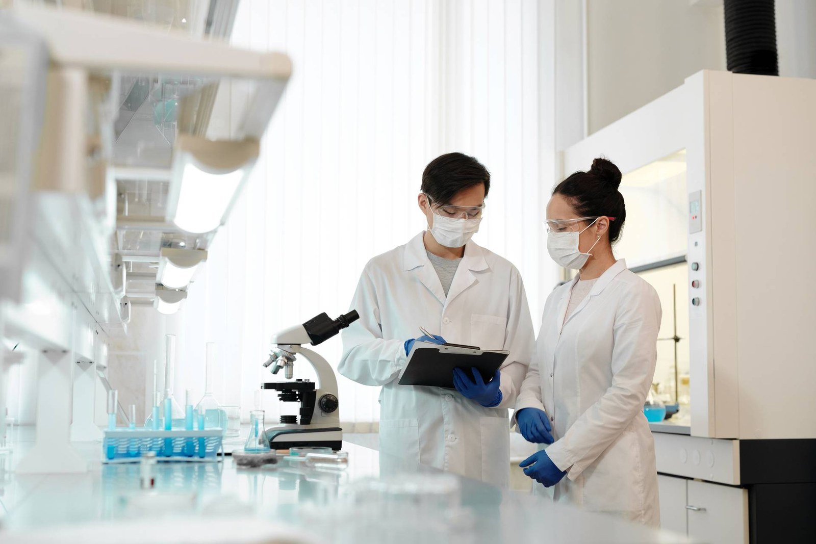 Two masked lab researchers in white coats and safety glasses review notes on a clipboard beside a microscope