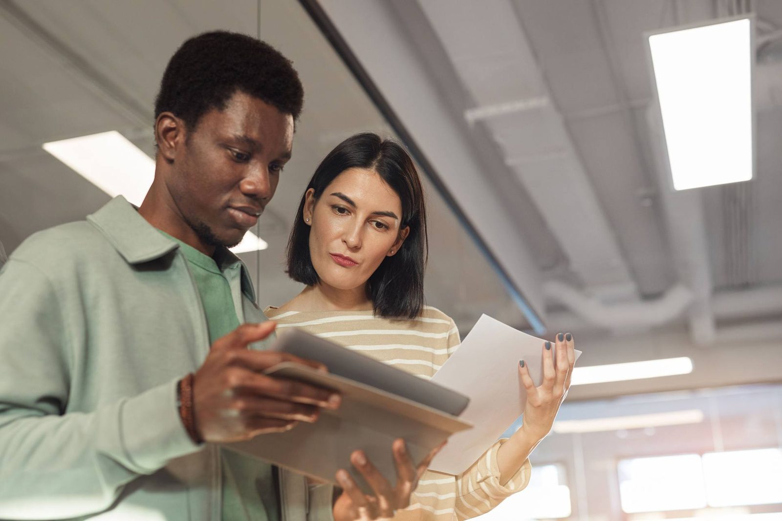 Two colleagues stand together reviewing a document and a tablet in a bright office