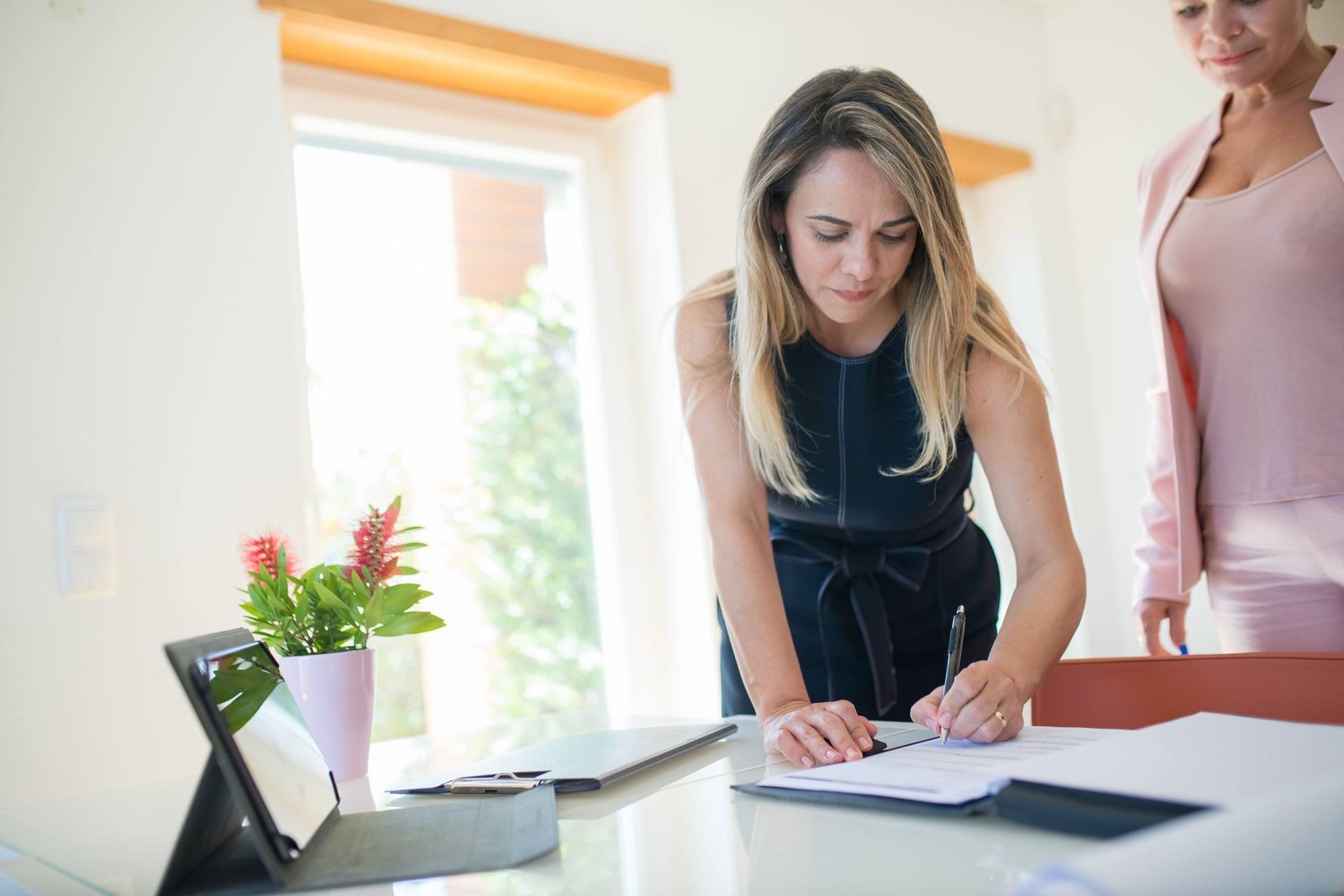 A woman leans over a desk signing an official document while a colleague looks on in a bright office