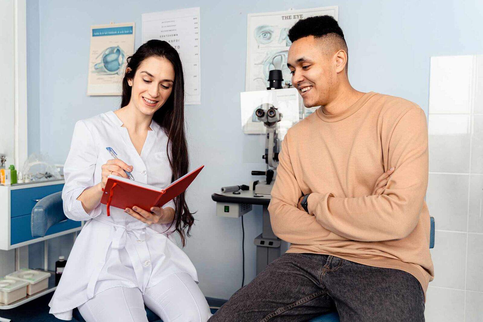 A clinician and patient smile together during an eye exam, with an eye-anatomy chart and slit lamp in the background