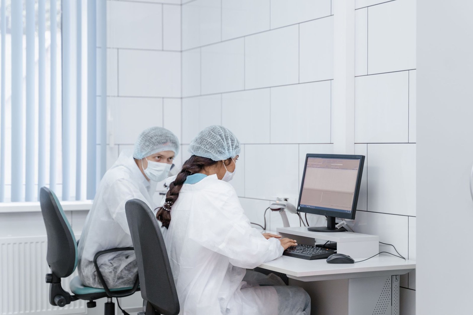 Two researchers in cleanroom coveralls, hairnets and masks review data on a computer monitor in a lab