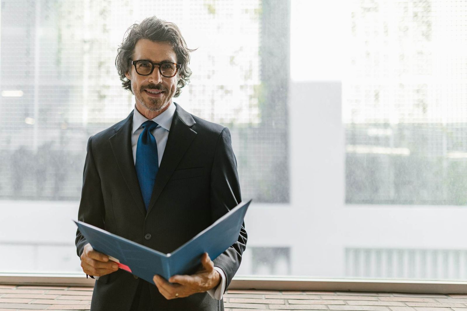 A smiling executive in a suit stands holding an open folder in a bright, window-lit space