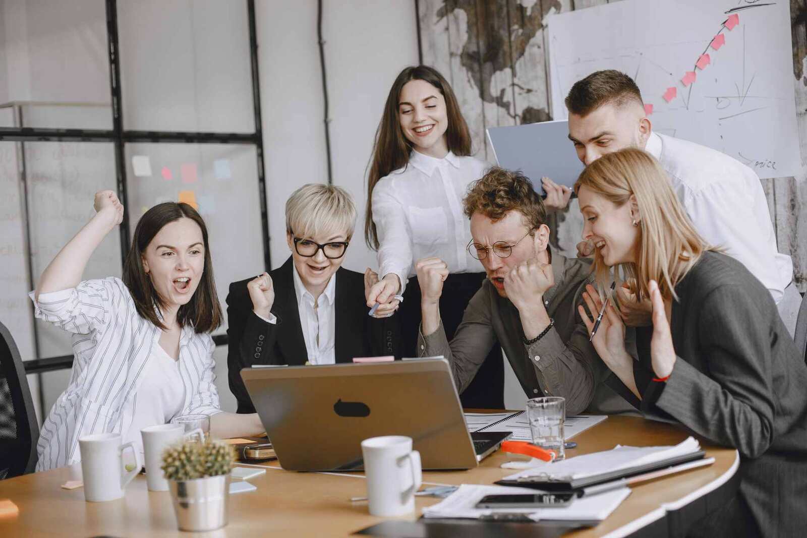 Six colleagues gather around a laptop in a bright office, pumping fists and cheering together