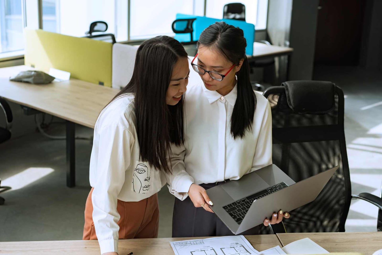 Two colleagues in a bright open office review a floor-plan drawing on a laptop and printed sheets