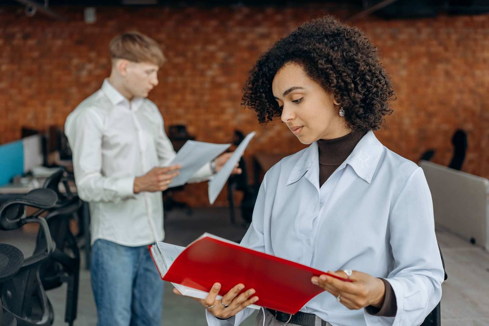 A woman holds an open red folder and reviews its contents while a colleague looks over papers behind her in a brick-walled office