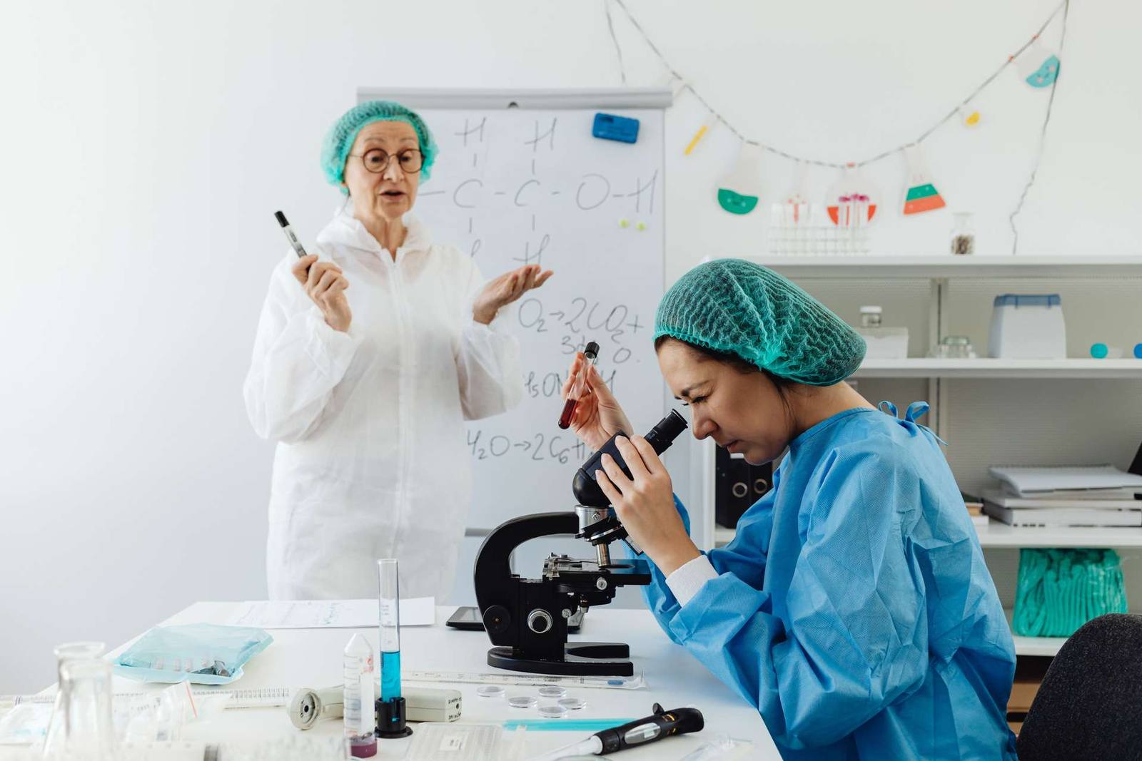 A researcher in a lab coat and hairnet gestures toward a whiteboard covered in chemical formulas while a colleague examines a sample under a microscope