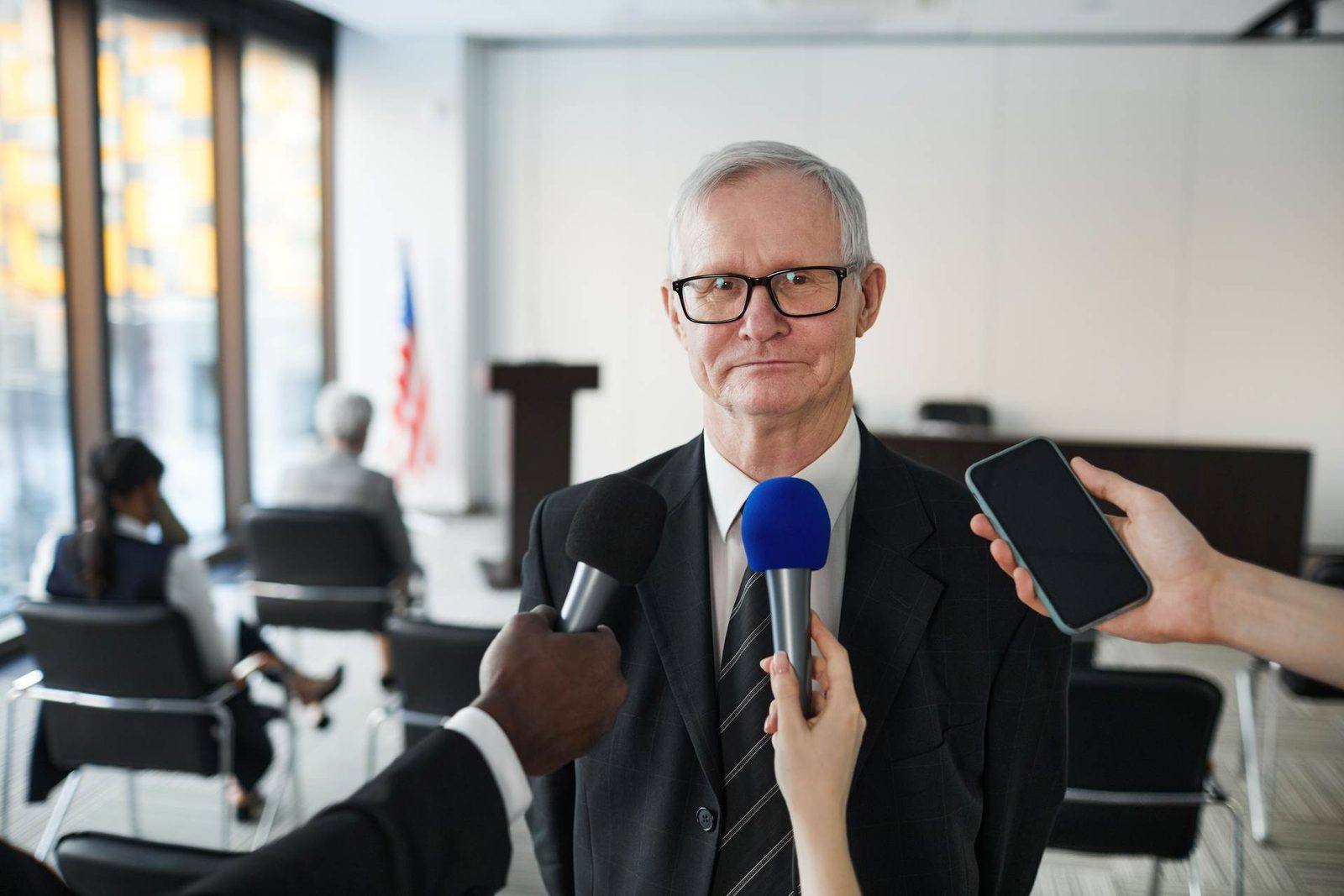 An older man in glasses and a suit stands answering questions as two hands hold microphones toward him and another hand holds up a phone to record