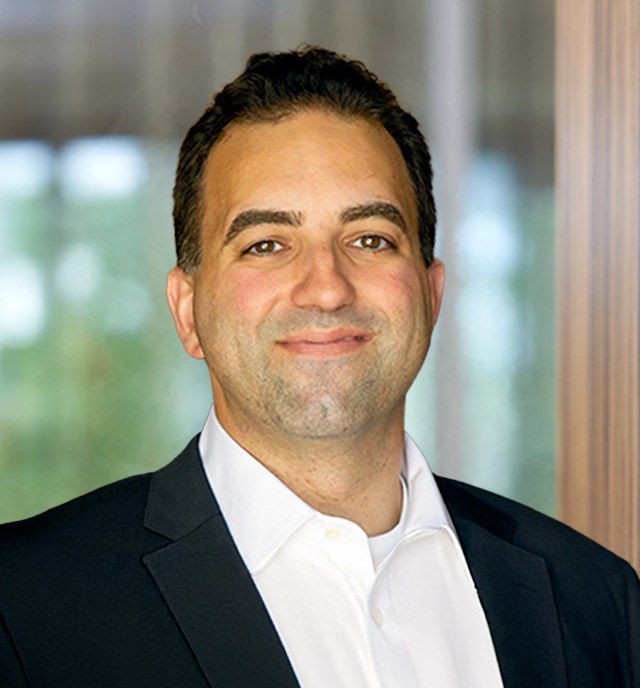 Head-and-shoulders portrait of Ryan Zeidan, a man with short dark hair in a dark blazer and white open-collar shirt, smiling in front of a blurred window