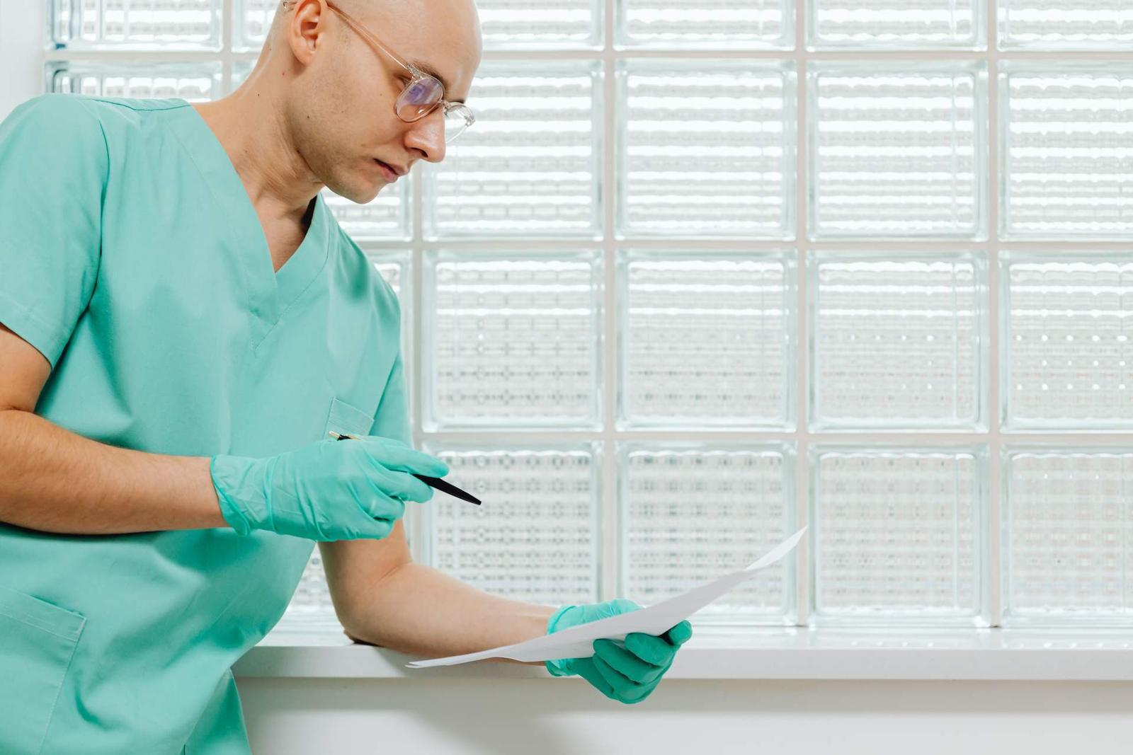 A gloved clinician in scrubs reviews a printed document by a bright glass-block window