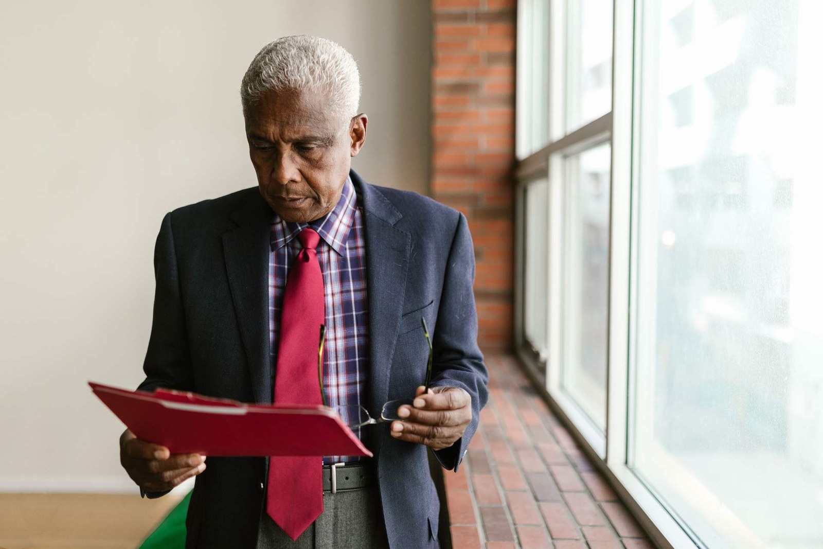 An executive reviews documents from a red folder while standing near a window
