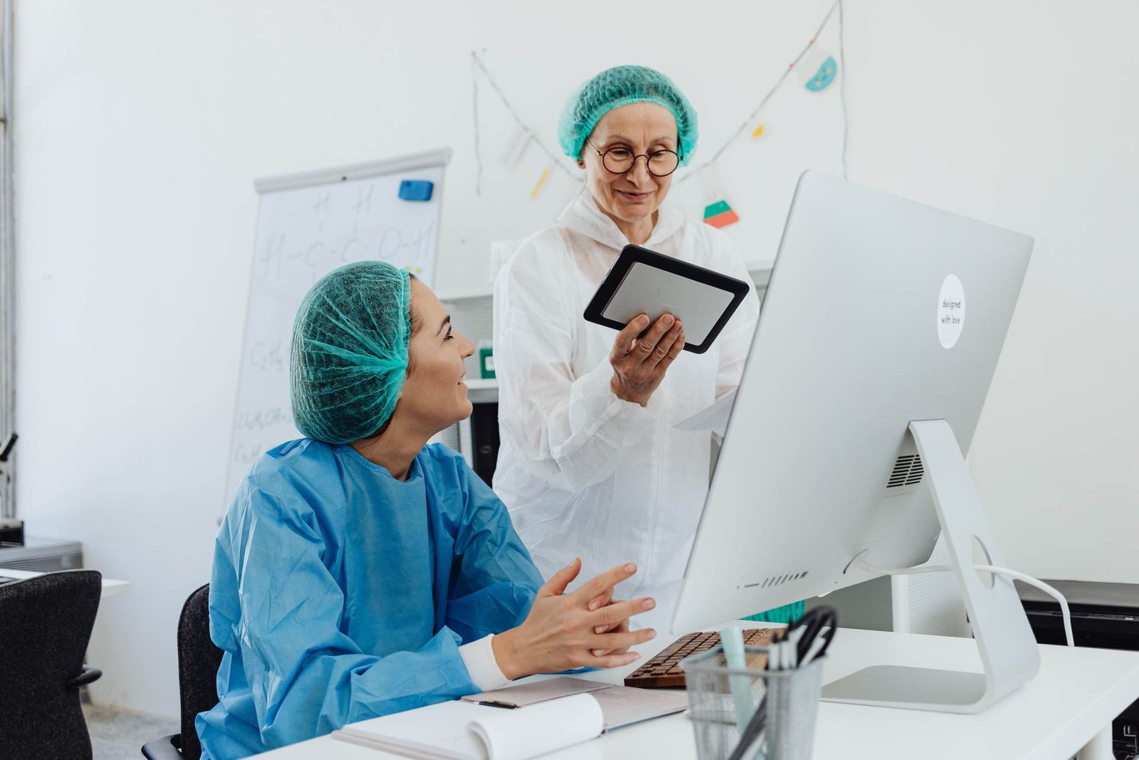 Two colleagues in surgical caps and protective gowns review notes on a tablet beside a desktop computer in a bright laboratory office
