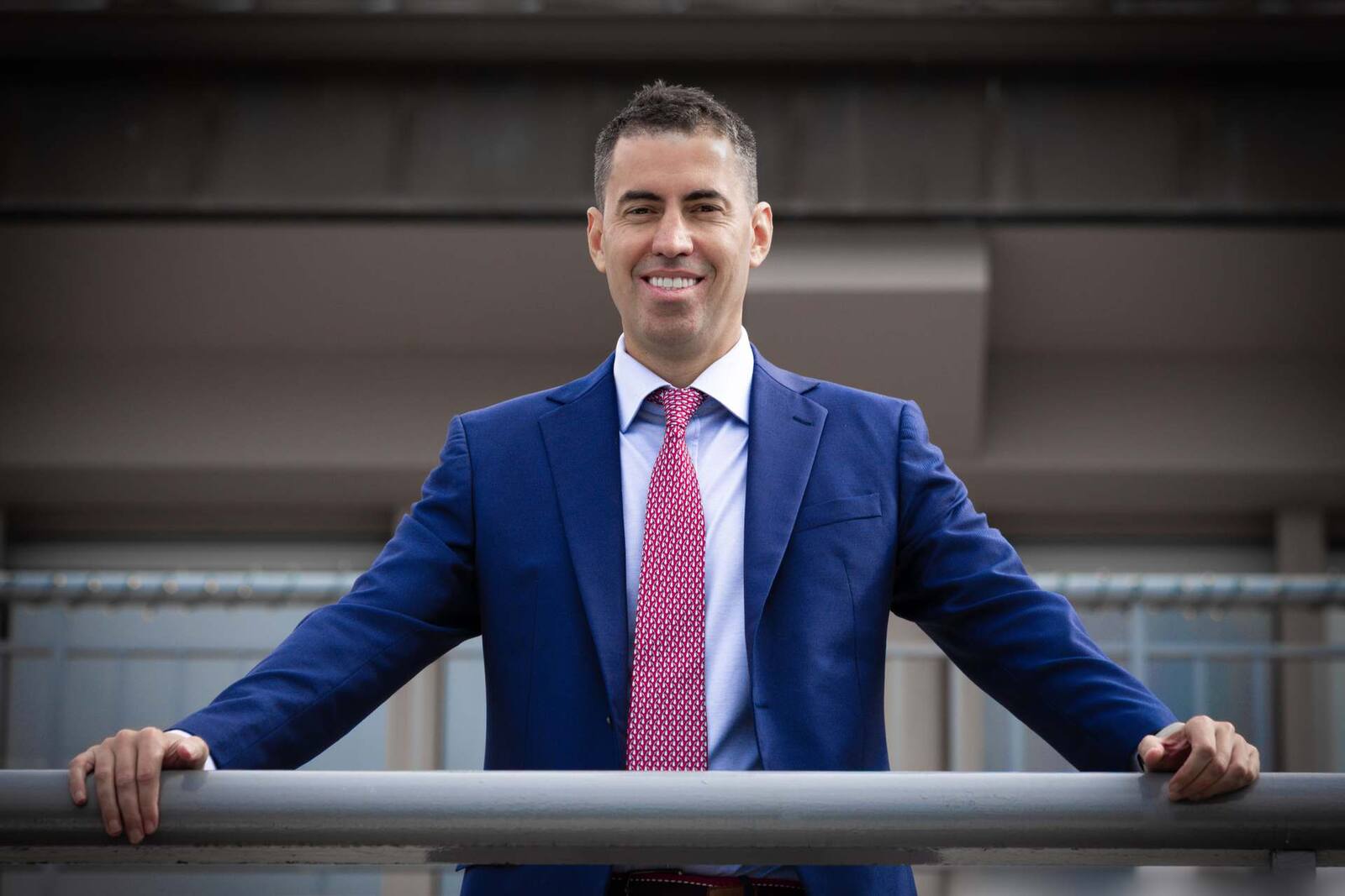 A smiling executive in a blue suit stands at an outdoor railing in front of an office building