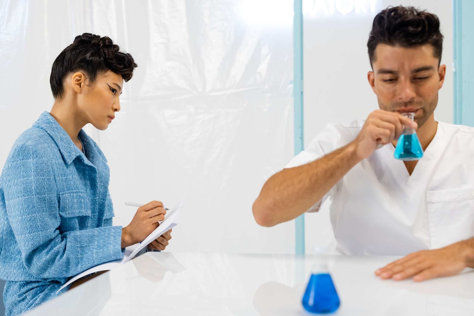 A researcher in white scrubs examines a conical flask of blue solution while a colleague in blue takes notes across the bench