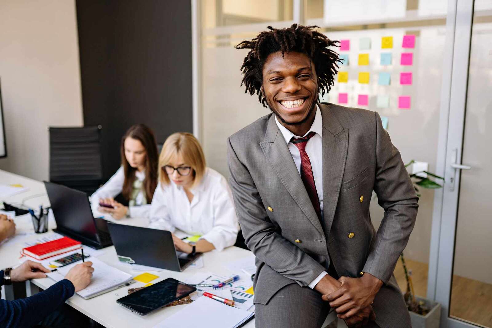 A smiling executive in a grey suit sits on the edge of a desk while two colleagues work at laptops behind him in a bright office