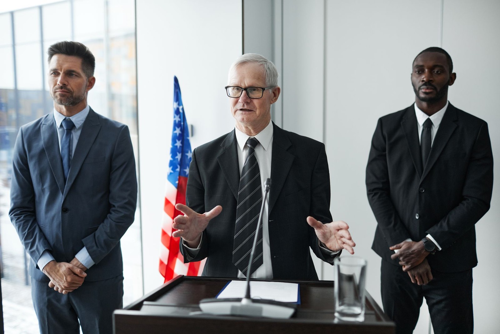 Three men in business suits stand at a lectern with a U.S. flag behind them, one gesturing as he speaks