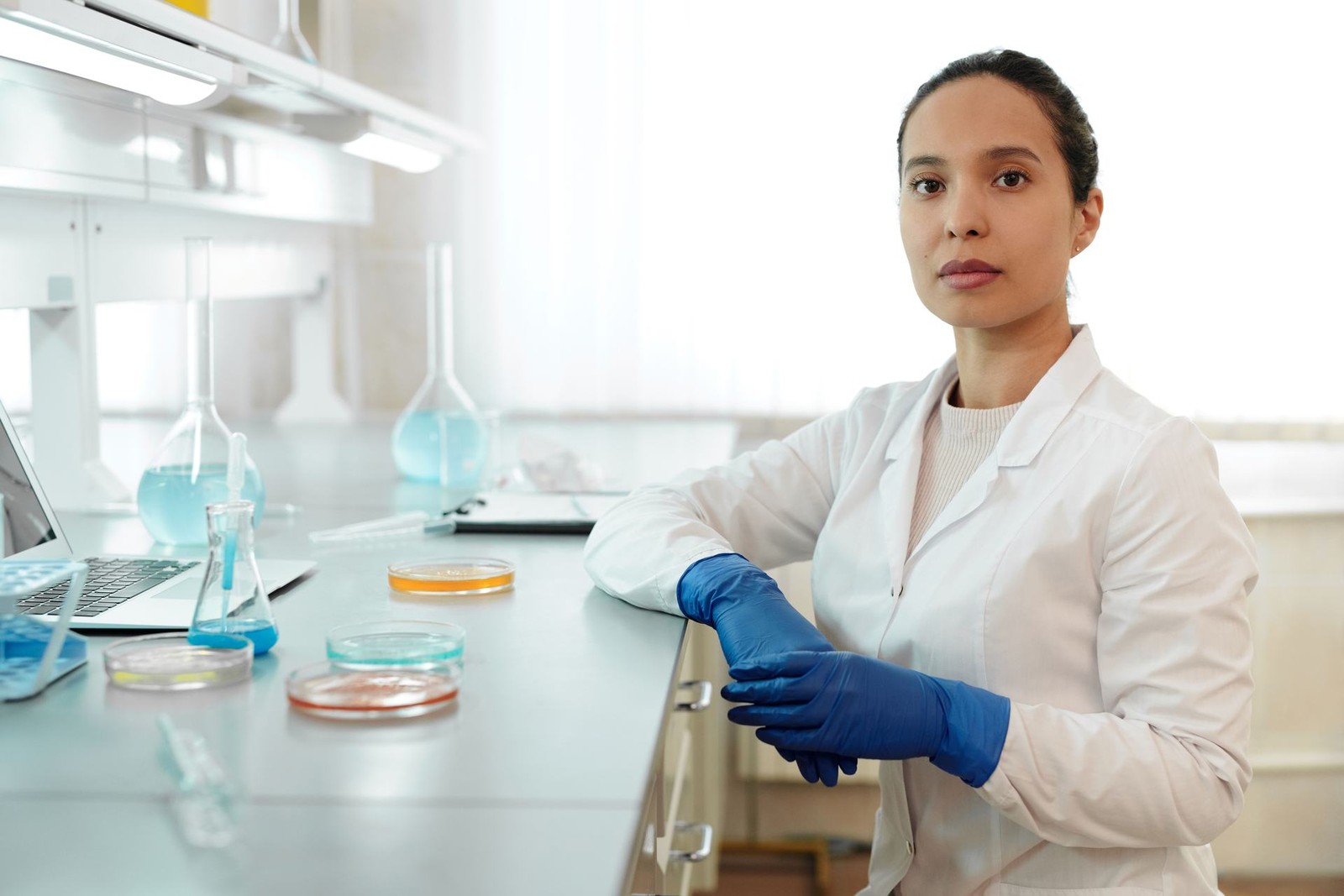 A scientist in a white lab coat and blue nitrile gloves stands at a laboratory bench beside petri dishes and glass flasks