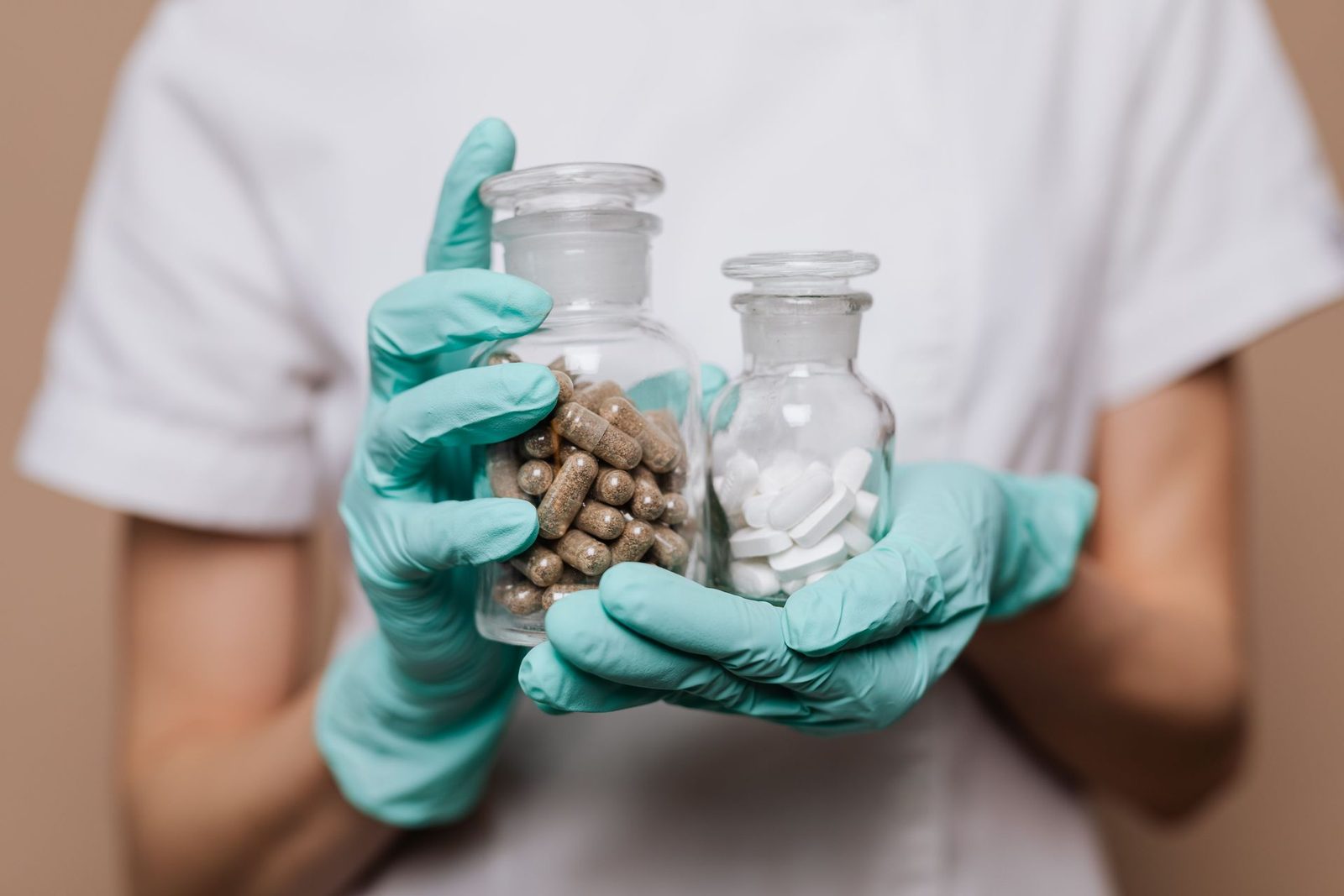 Gloved hands hold two apothecary jars, one filled with capsules and one with tablets