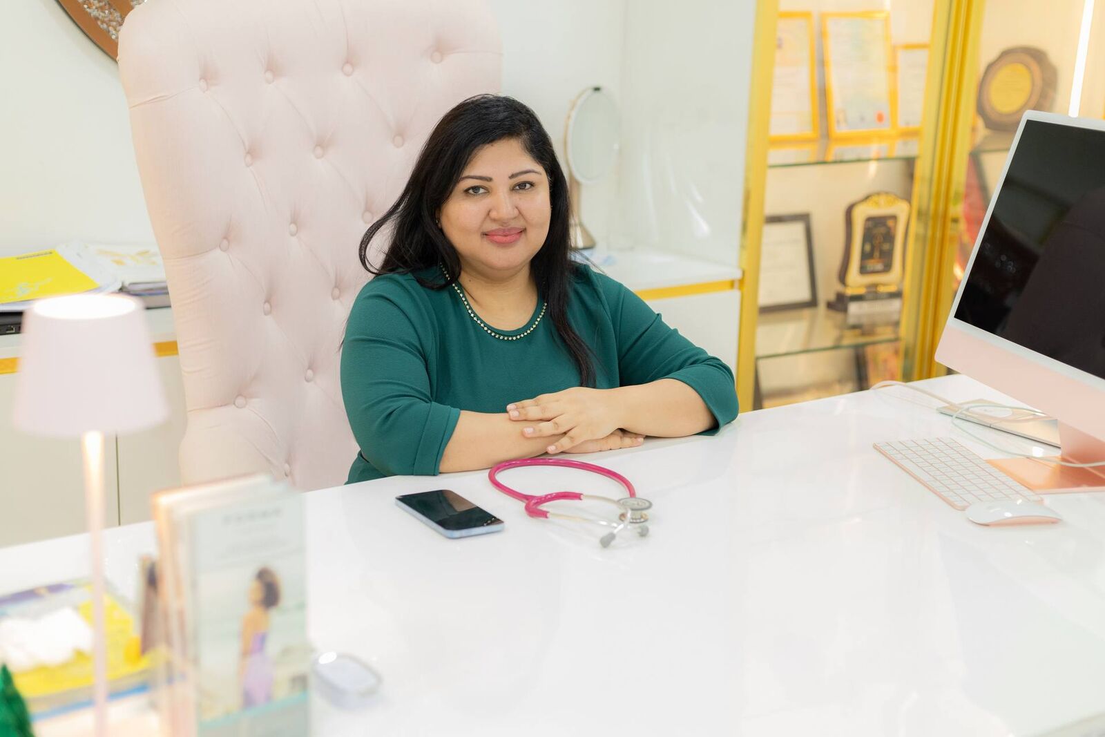 A physician sits at a bright, tidy desk with a stethoscope, a phone and a desktop computer