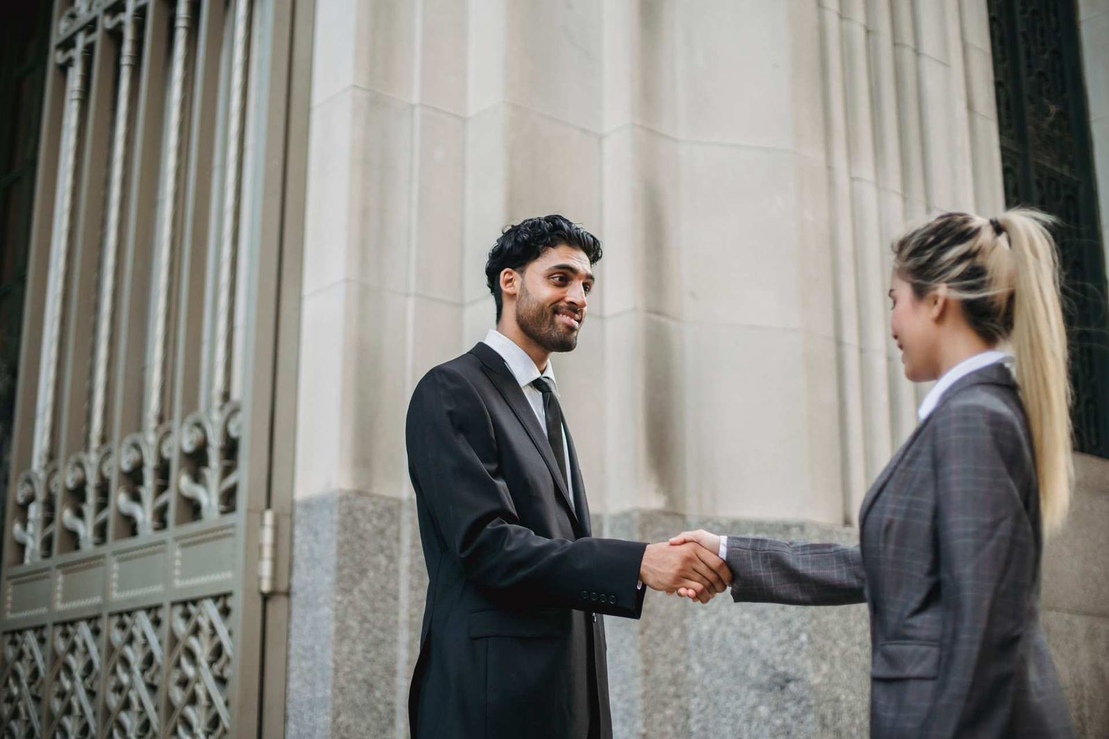 Two professionals in suits shake hands in front of a stone-columned government building facade