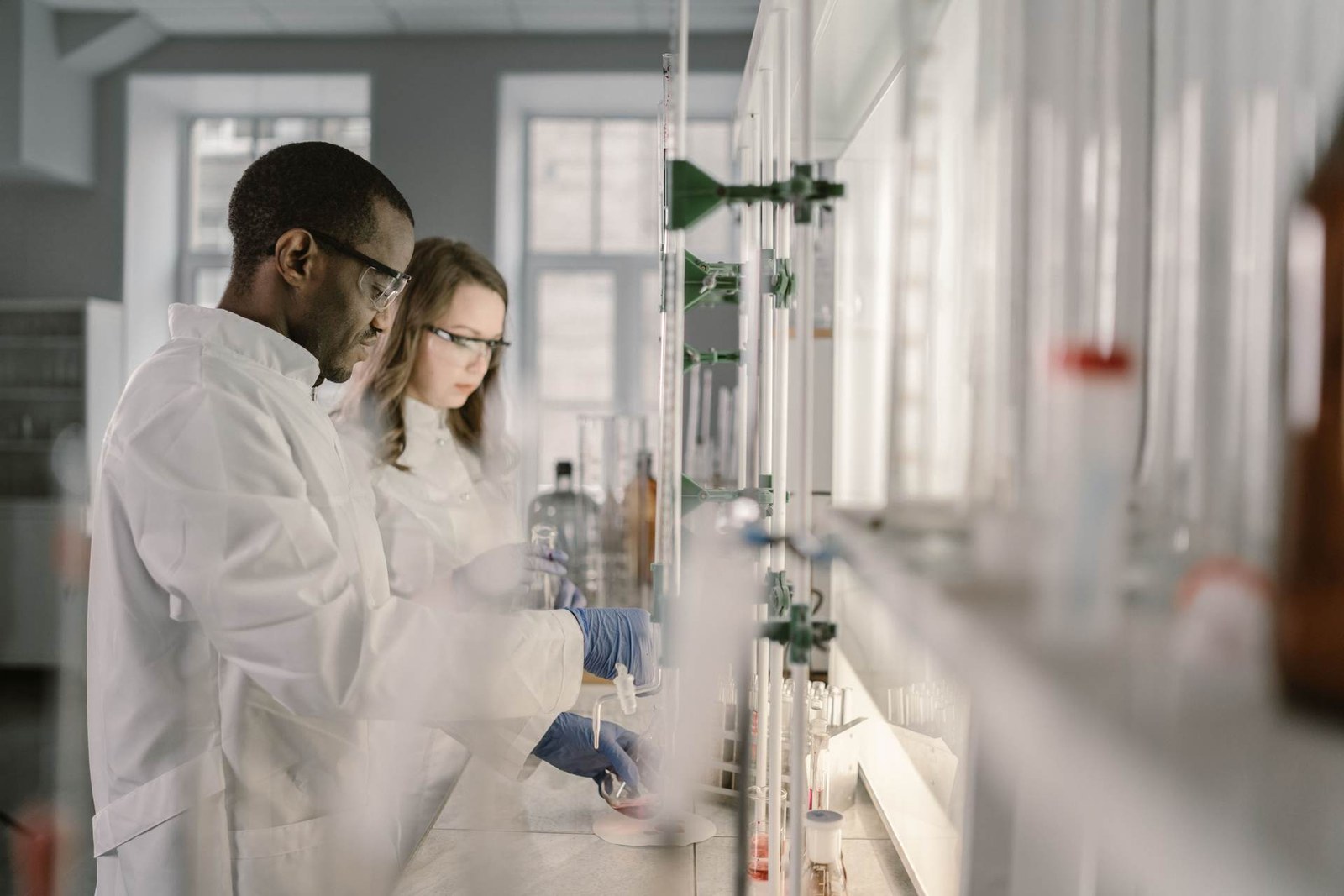 Two lab scientists in white coats test glassware samples on a bright laboratory bench
