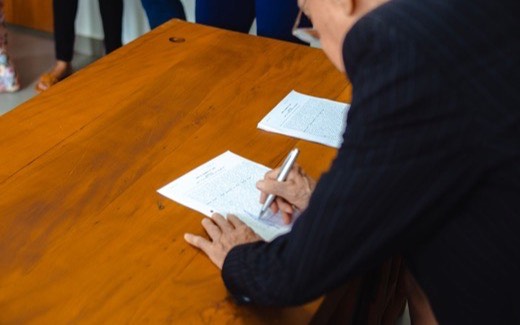 A person in a dark jacket signs printed documents at a wooden desk while colleagues stand nearby