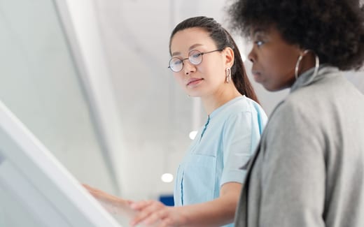 A clinician in a light blue coat points to something on a bright wall-mounted screen while a colleague looks on