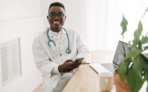 A clinician in a white coat smiling while checking a phone beside an open laptop in a bright office