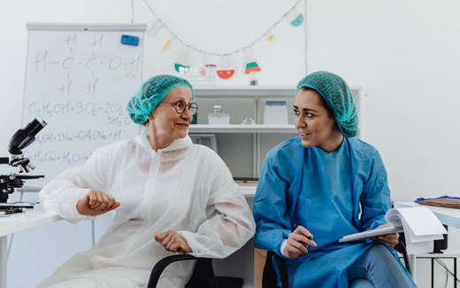 Two researchers in cleanroom hairnets and gowns talk beside a whiteboard covered in chemistry notation