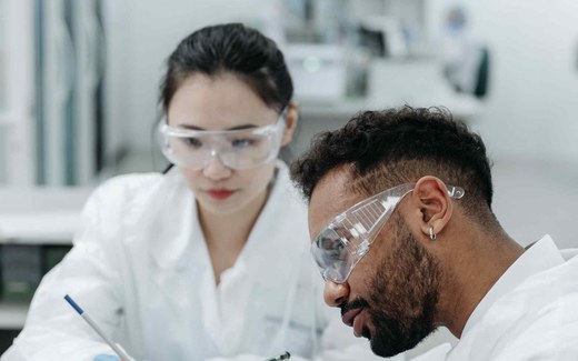 Two lab specialists in white coats and safety glasses examine a blood-collection tube in a clinical laboratory