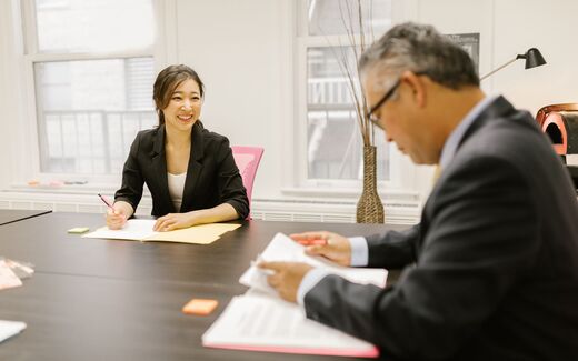 Two colleagues review and sign paperwork together at an office table
