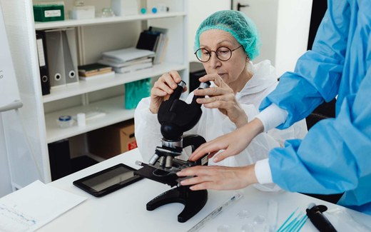 A researcher in a lab coat adjusts a microscope while a colleague in scrubs assists, examining a sample under magnification