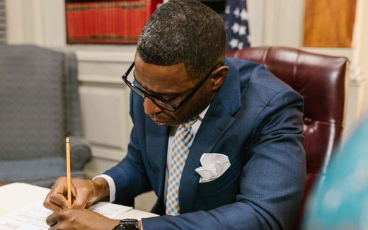 A man in a suit reviews and signs a paper document at a desk, with an American flag in the background