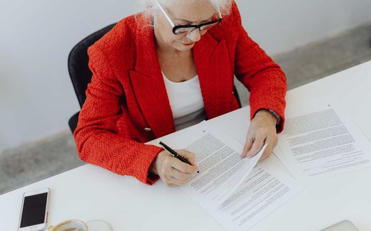 A woman in a red blazer reviews and signs printed documents at a white desk