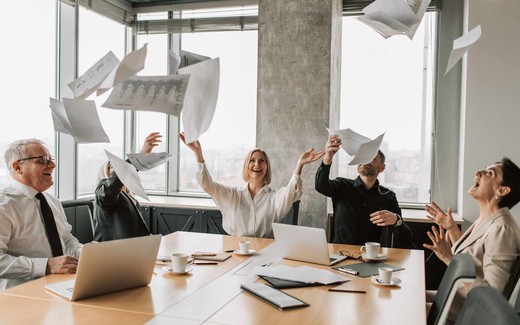 A team of colleagues in a bright office celebrate a milestone, tossing papers into the air around a conference table