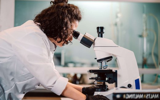 A researcher in a white lab coat looks into a benchtop microscope
