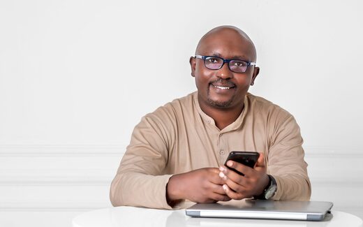 A smiling professional in business attire sits at a white table, glasses on, holding a phone beside a closed laptop