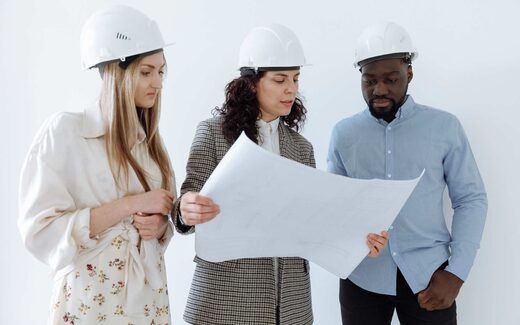 Three professionals in hard hats reviewing facility plans, a generic stand-in for a manufacturing-site inspection or remediation review