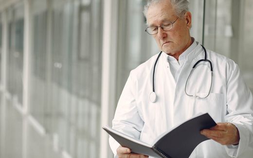 A senior physician in a white coat reads clinical notes in a hospital corridor