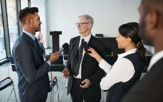 An executive in a dark suit takes questions from two reporters in a bright office