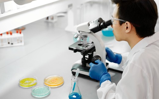 A researcher in a lab coat examines petri dish cultures under a microscope