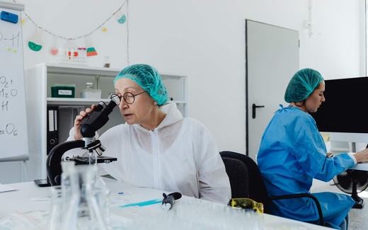A researcher in a lab coat and hairnet looks into a microscope at a bench lined with glassware, with a colleague working at a computer behind her